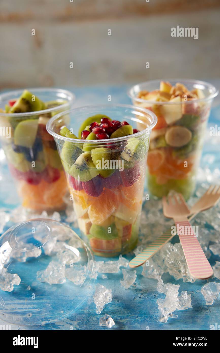 Mezcla de frutas frescas y sabrosas picadas en vasos de plástico Coloque la mesa con hielo y tenedores de madera en una habitación luminosa Foto de stock