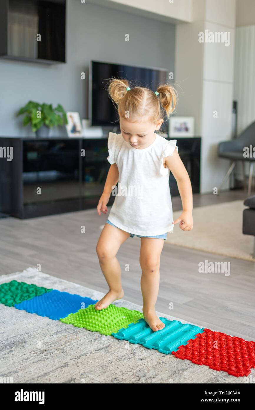 Una niña pequeña camina sobre una alfombra de masaje. Alfombrilla de masaje de pies para bebés. Ejercicios para piernas alfombra de masaje ortopédico. Foto de stock
