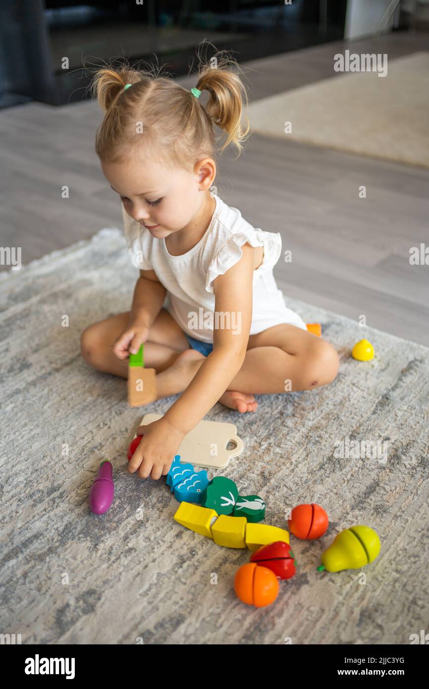 Linda niña pequeña jugando en casa con juguetes de madera ecológicos. Niño feliz cortando verduras y frutas con cuchillo de juguete. El niño jugando educativo Foto de stock