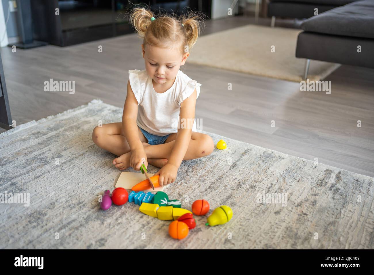 Linda niña pequeña jugando en casa con juguetes de madera ecológicos. Niño feliz cortando verduras y frutas con cuchillo de juguete. El niño jugando educativo Foto de stock