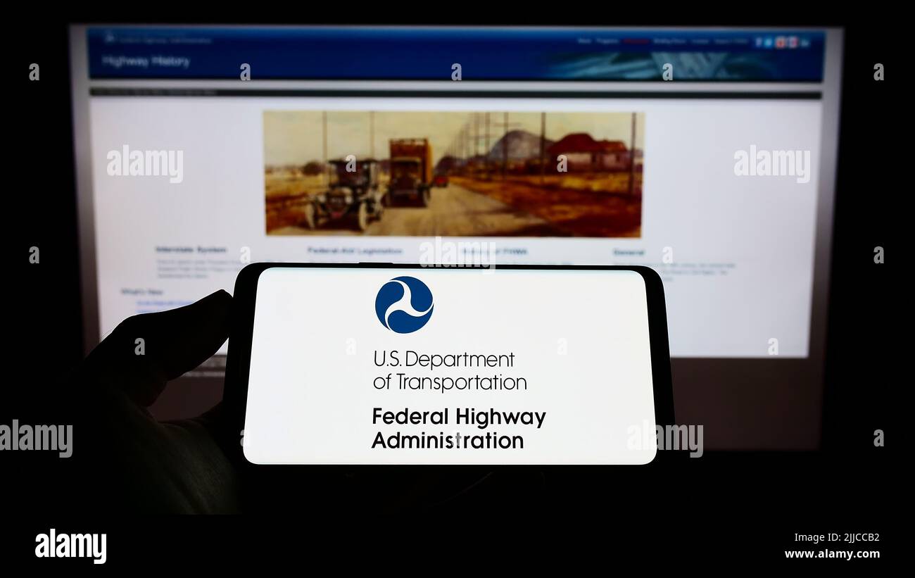 Persona que sostiene el teléfono celular con el logotipo de la Administración Federal de Carreteras de América (FHWA) en la pantalla delante de la página web. Enfoque la pantalla del teléfono. Foto de stock