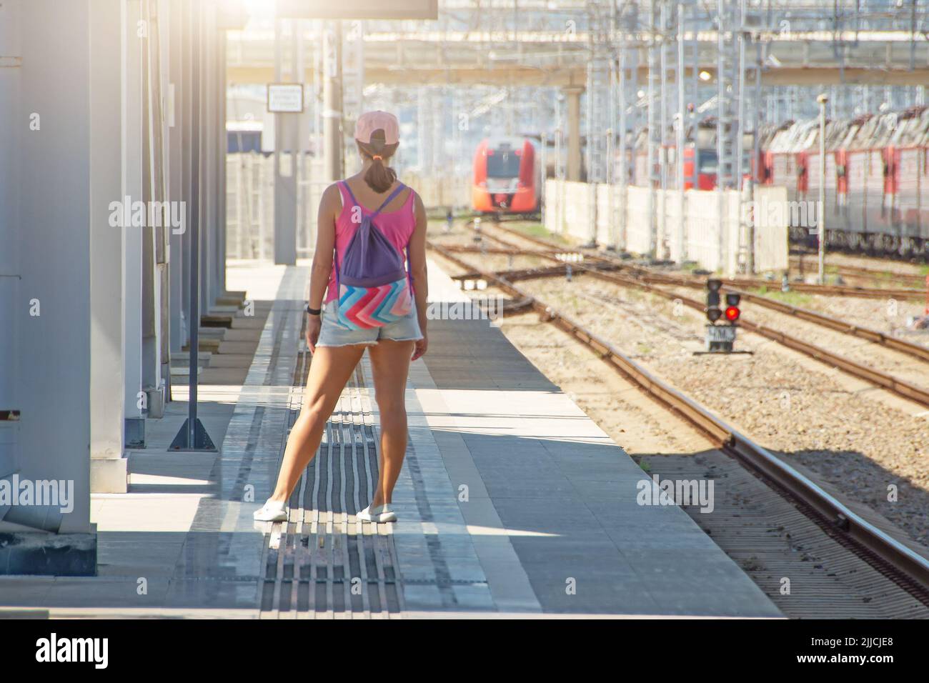 Una chica con ropa de verano y una mochila esperando a que llegue un tren eléctrico de cercanías a la distancia Foto de stock