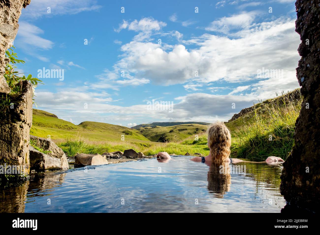 Mujer relajarse y bañarse en la piscina natural geotérmica caliente, aguas termales en Islandia en verano. Verdes colinas ondulantes y cielo azul en el fondo. Caliente Foto de stock