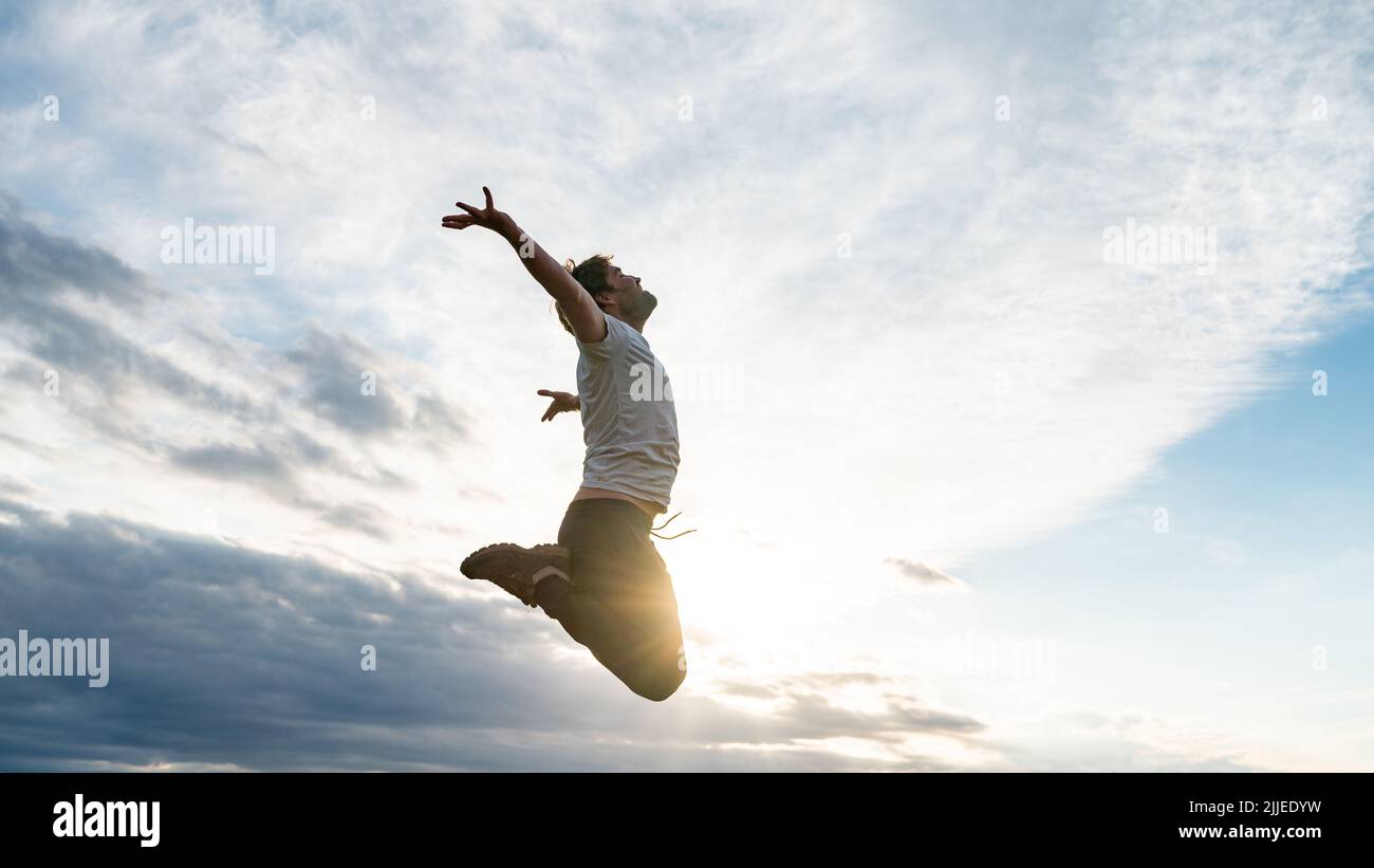 Hombre joven emocionado con ropa informal saltando en lo alto del aire, inclinando sus brazos hacia el cielo retroiluminado por el sol de la tarde. Foto de stock