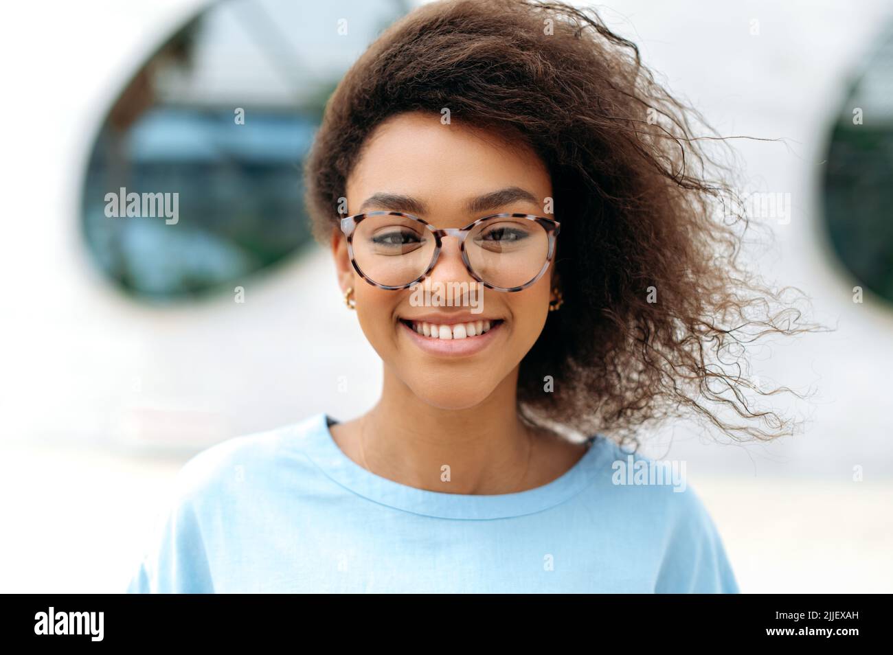 La foto de primer plano de una mujer de negocios o estudiante afroamericana encantadora positiva y segura de pelo rizado, con gafas, de pie al aire libre, mira la cámara con una sonrisa feliz y bonita Foto de stock