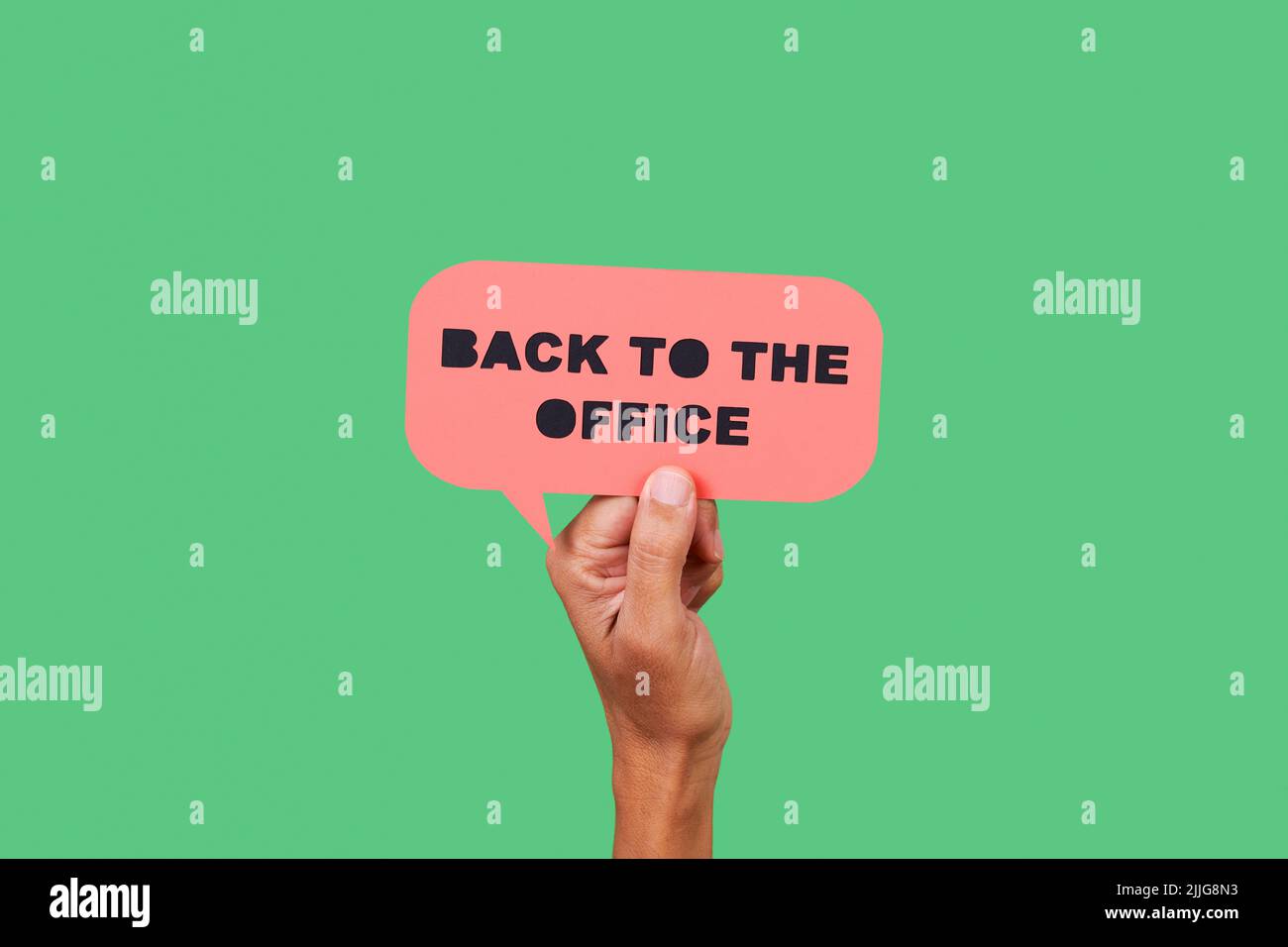 primer plano de la mano de un hombre sosteniendo una burbuja de discurso de papel rosa con el texto de vuelta a la oficina en ella, sobre un fondo verde Foto de stock
