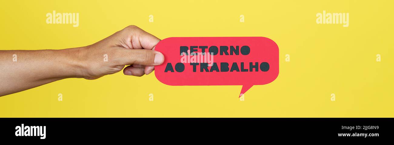 la mano de un hombre sosteniendo un cartel de papel rojo con el texto de vuelta a trabajar escrito en portugués, sobre un fondo amarillo, en un formato panorámico para usar como w Foto de stock