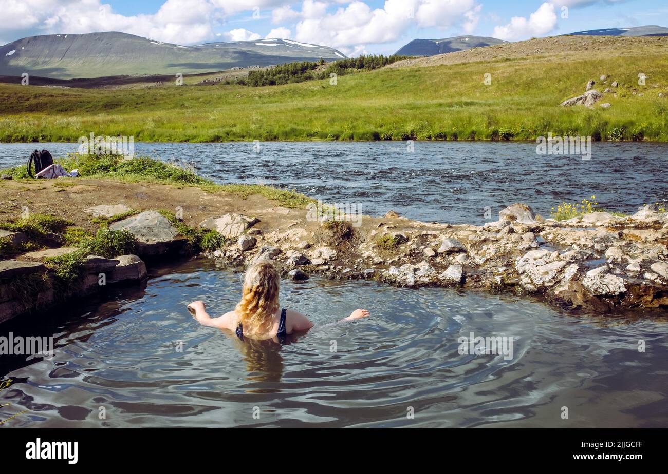 Mujer relajarse y bañarse en la piscina natural geotérmica caliente, aguas termales en Islandia en verano. Verdes colinas ondulantes y cielo azul en el fondo. Foto de stock