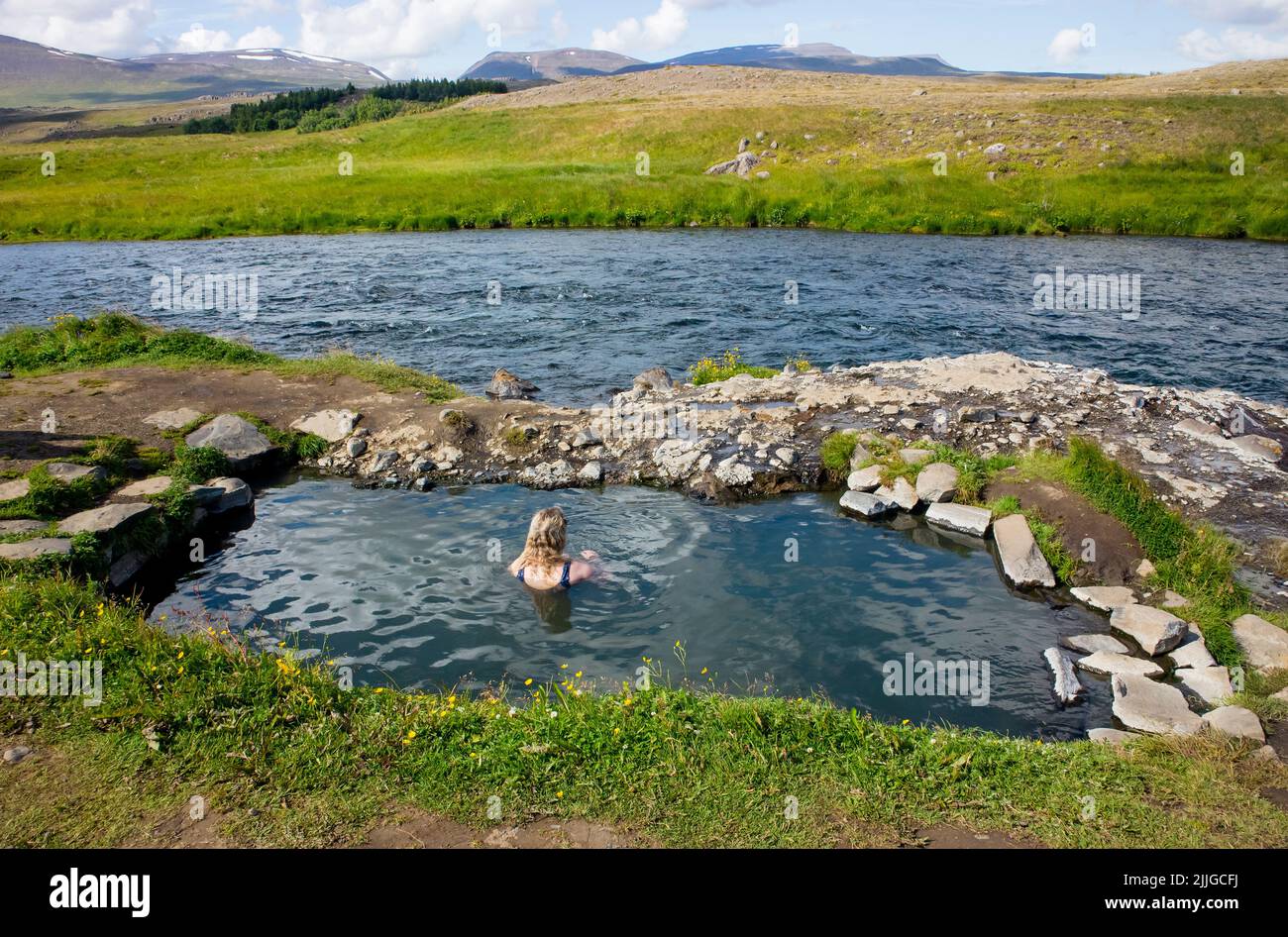 Mujer relajarse y bañarse en la piscina natural geotérmica caliente, aguas termales en Islandia en verano. Verdes colinas ondulantes y cielo azul en el fondo. Foto de stock
