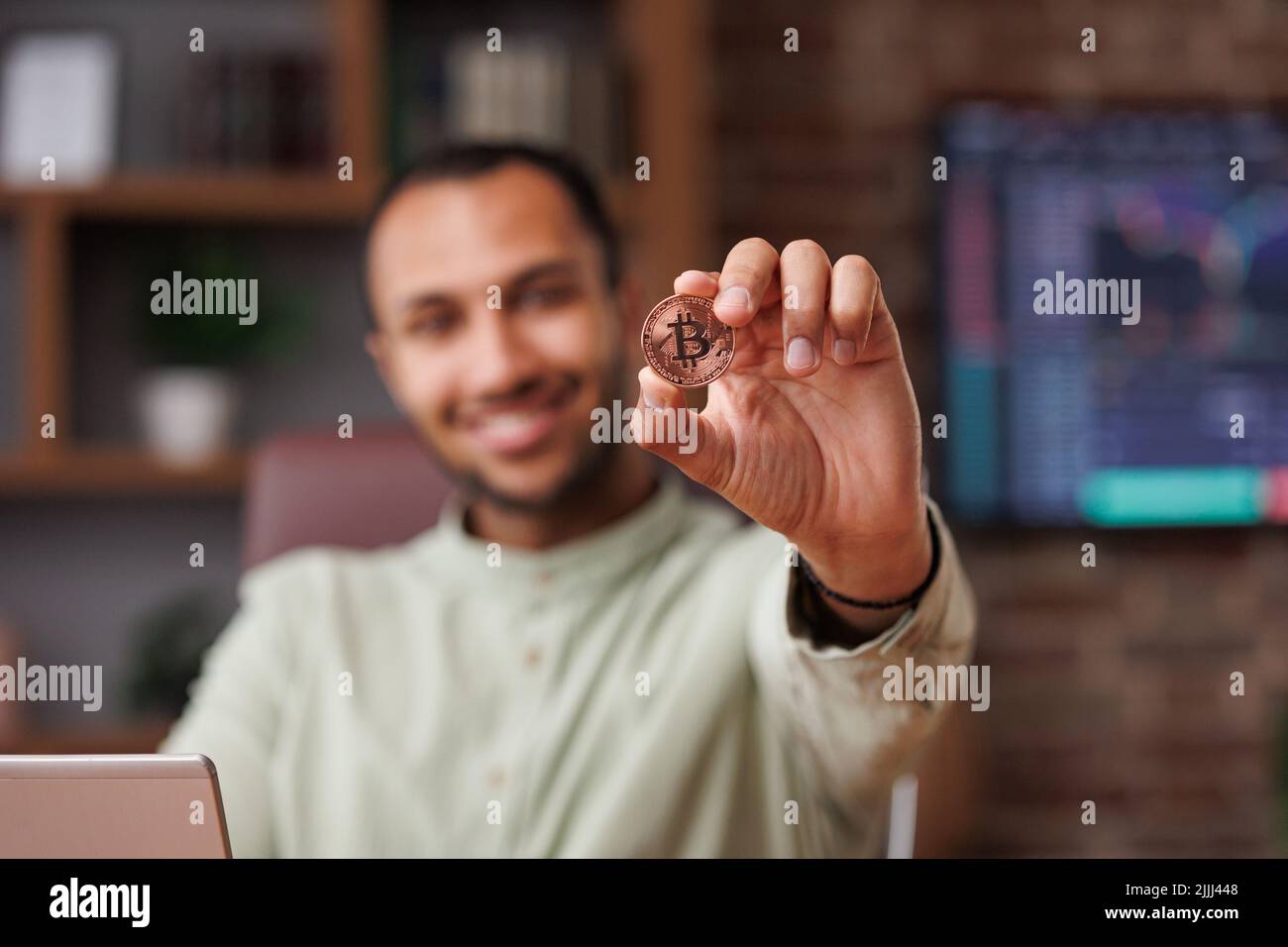 Retrato de un sonriente hombre de negocios sosteniendo bitcoin en el interior de la oficina con gráficos de la bolsa en el fondo. Usando bitcoins como pago. Investigaciones en Foto de stock