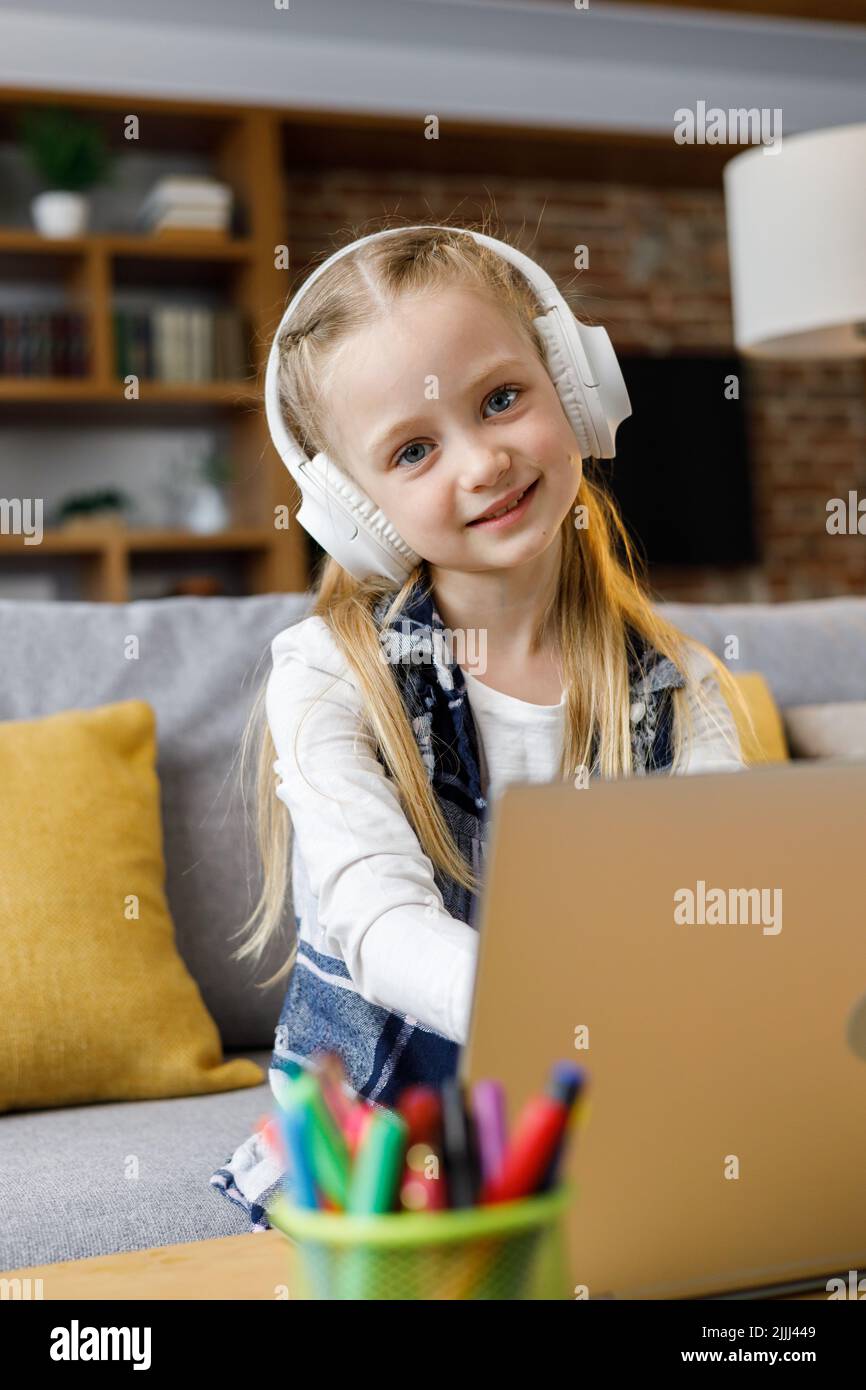 Retrato de una linda niña de escuela primaria estudiando en casa usando una computadora portátil. Niño sonriente con auriculares blancos trabajando en el ordenador. Distancia Foto de stock