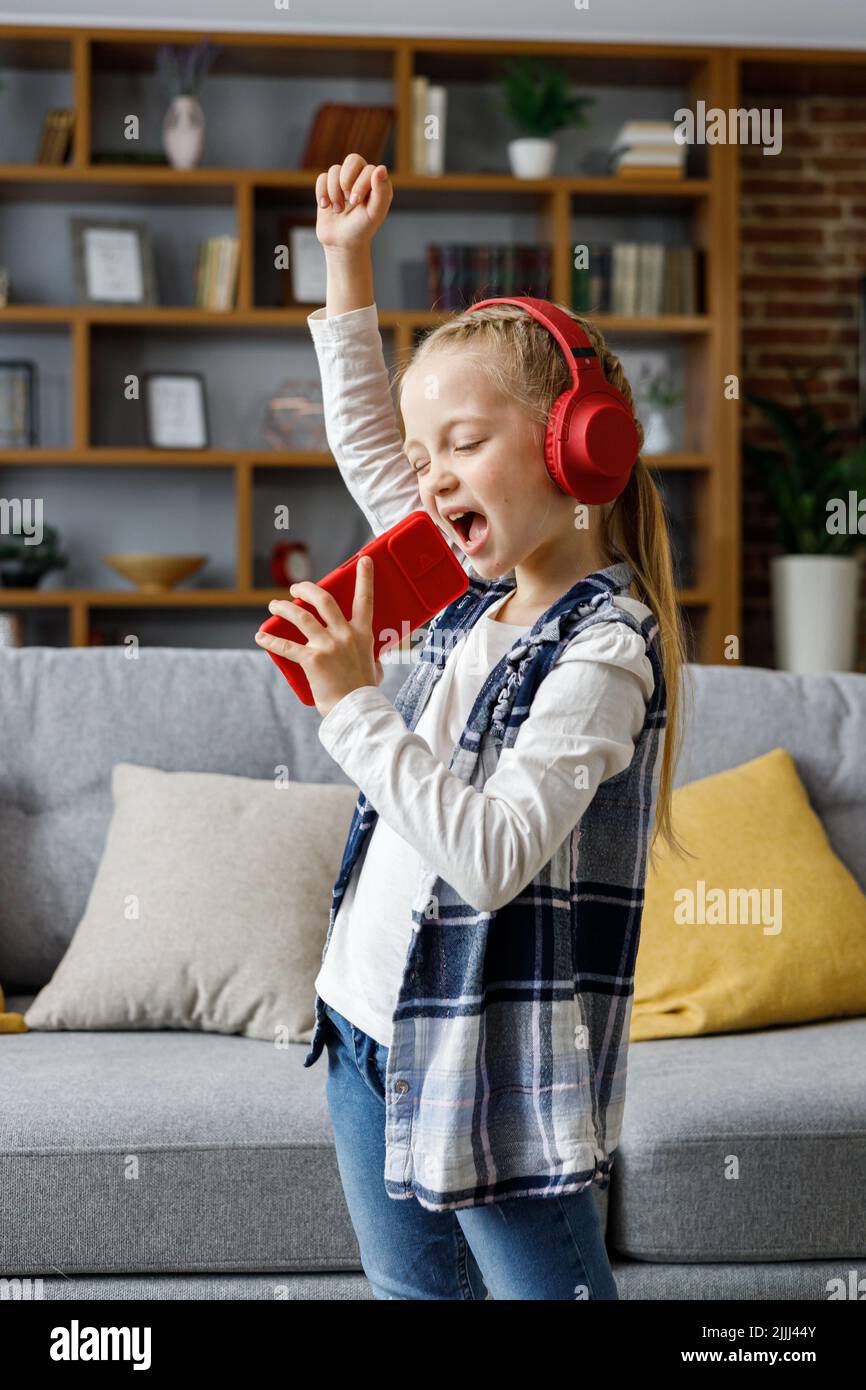 Niña feliz con auriculares rojos bailando y cantando con micrófono como un smartphone. Niño lindo disfrutar escuchando sus pistas de música favoritas en Foto de stock