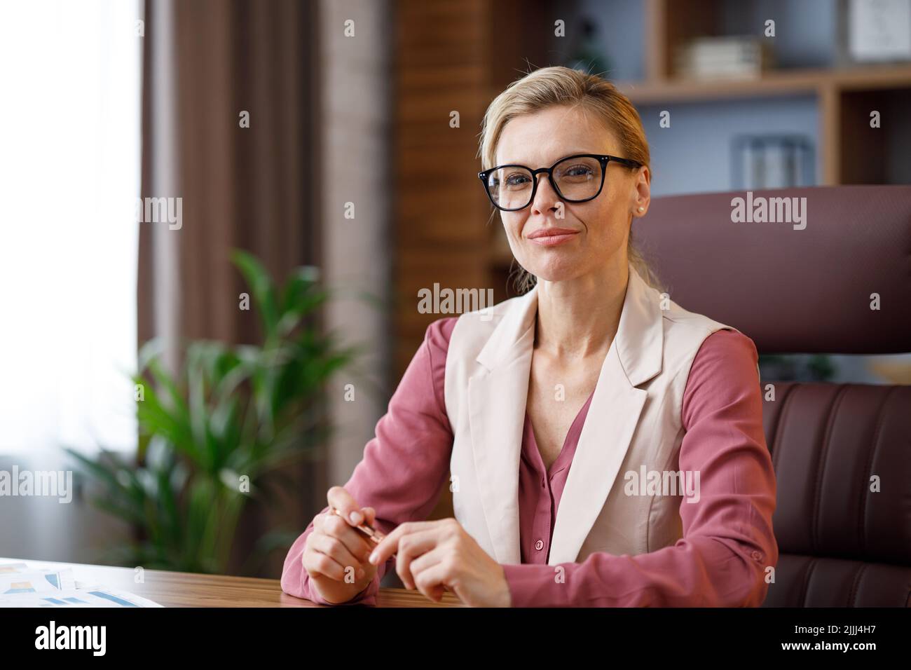 Retrato de la hermosa mujer blondie mirando la cámara y sonriendo. Mujer de negocios segura y con estilo para adultos con gafas sentado en el lugar de trabajo Foto de stock