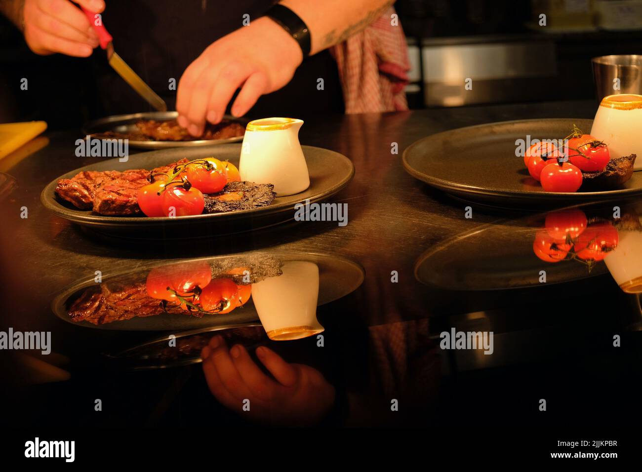 Las manos del chef preparan un plato de comida para los clientes en una cocina comercial. Bistec de ternera, tomates de vid, champiñones, salsa de pimienta presentada en el plato. Foto de stock