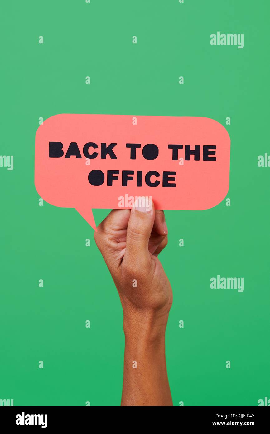el hombre sostiene un cartel de papel rosa, en forma de burbuja de discurso, con el texto de vuelta a la oficina en él, sobre un fondo verde Foto de stock