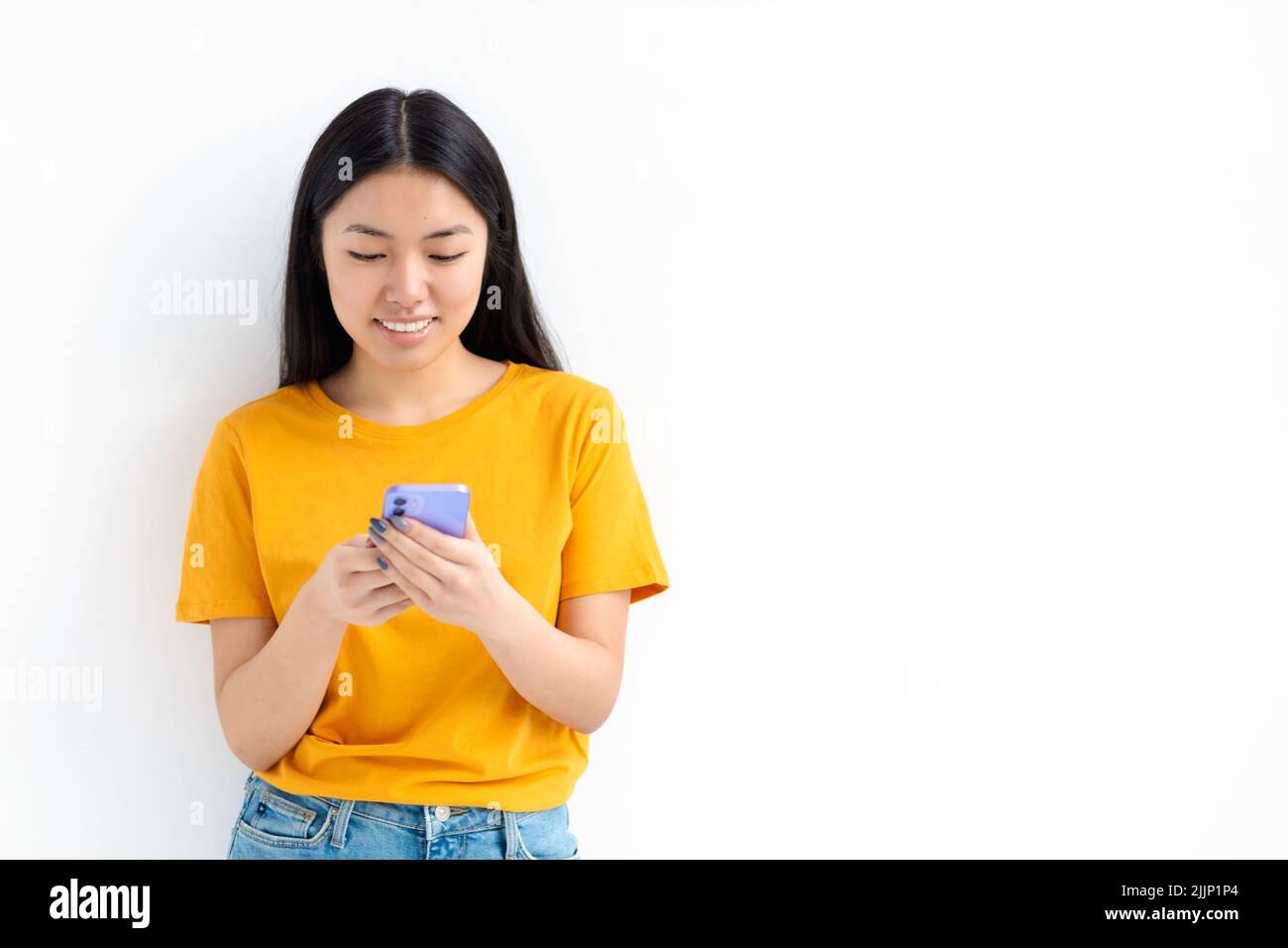 Hermosa joven alegre mujer asiática utilizando el teléfono móvil y sonriendo de pie sobre fondo blanco. Chateando en línea Foto de stock