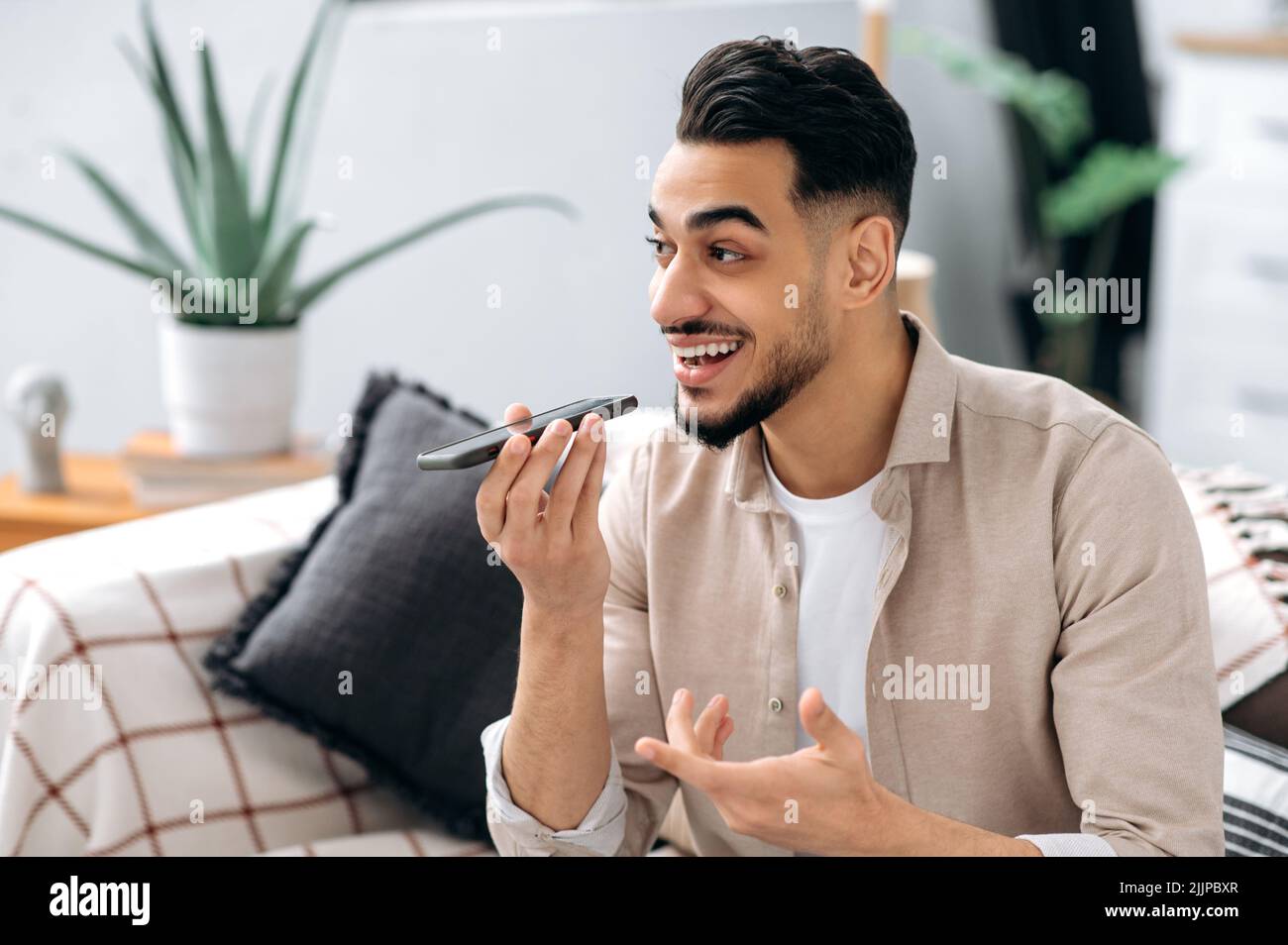 Un hombre indio o árabe emocionado, sentado en un sofá de la sala de estar, sosteniendo su smartphone, hablando con el altavoz y sonriendo, graba un mensaje de voz. Concepto de tecnología inalámbrica Foto de stock