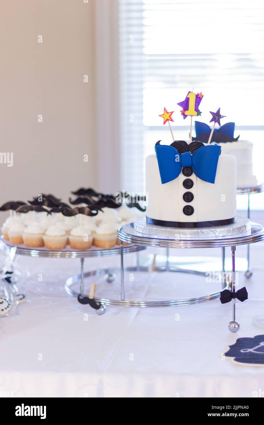 Un pequeño pastel de cumpleaños con pajarita azul y pequeños cupcakes a un lado Foto de stock