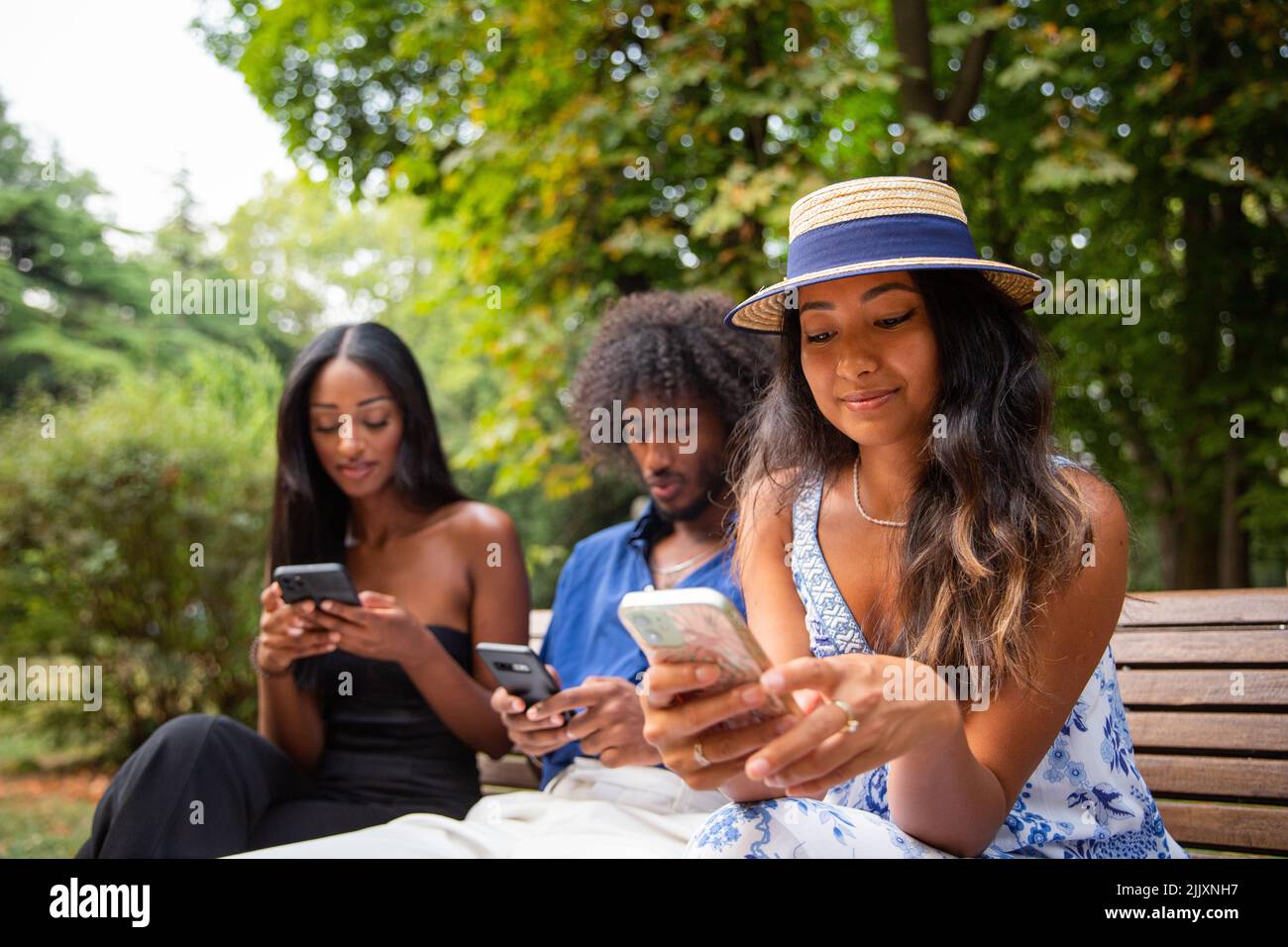 Tres amigos sentados en un banco en un parque utilizan sus smartphones, grupo multiétnico. Foto de stock