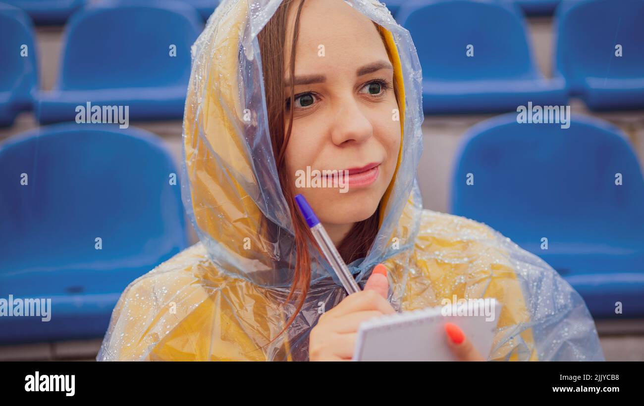 Mujer joven en impermeable con bloc de notas, bolígrafo sentado en gradas del estadio solo en tiempo lluvioso. Periodista escribiendo notas durante el trai deportivo Foto de stock