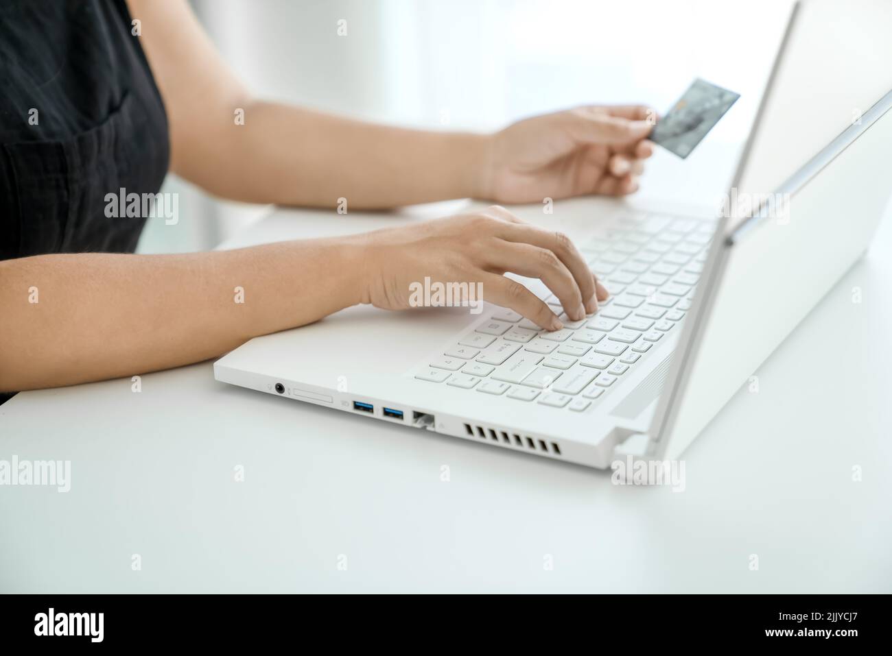 Mujer joven hace compras en línea sentado frente al ordenador portátil con la tarjeta bancaria en la mano. Manos en primer plano. Concepto de compras en línea y transferencia de dinero. Foto de stock