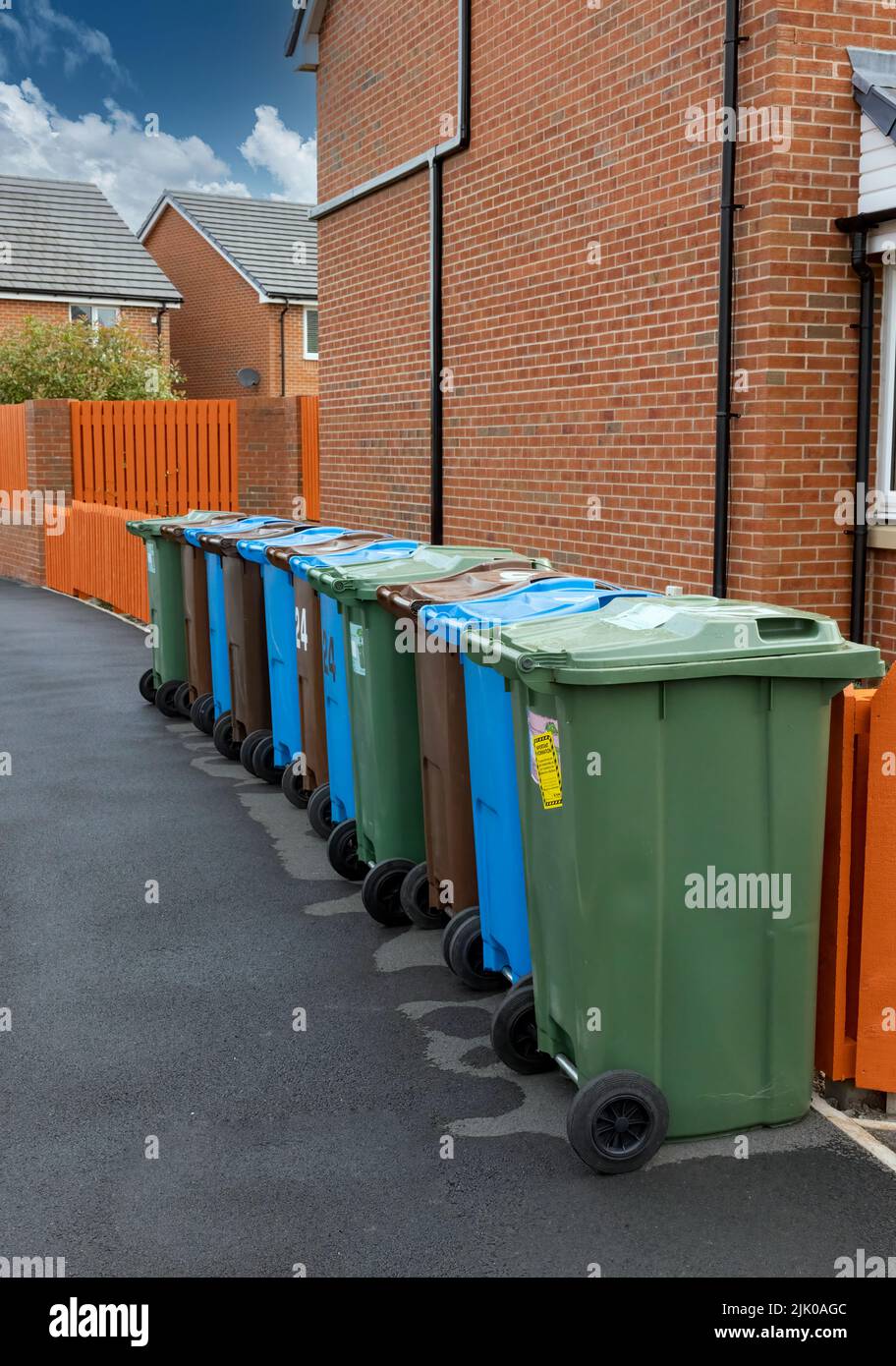 Una fila de contenedores de ruedas de colores para reciclar plástico, papel y residuos de jardín, junto a una cerca naranja y fuera de una casa moderna. Foto de stock