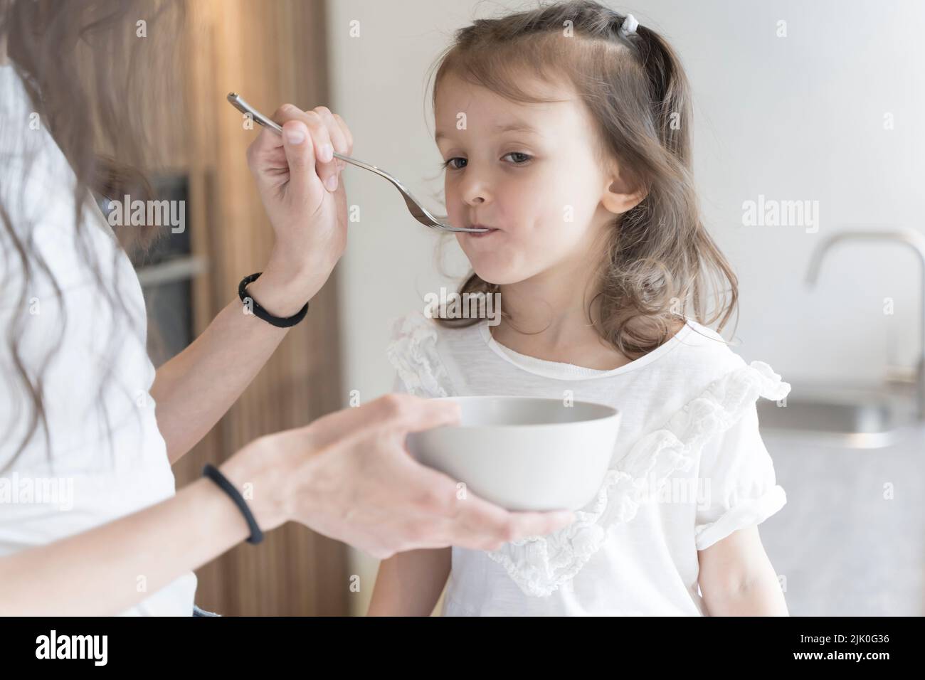 Mamá alimenta a su hija el desayuno en la cocina de la casa por la mañana. Concepto de comida para bebés, familia, cuidado. Fotografía de alta calidad Foto de stock