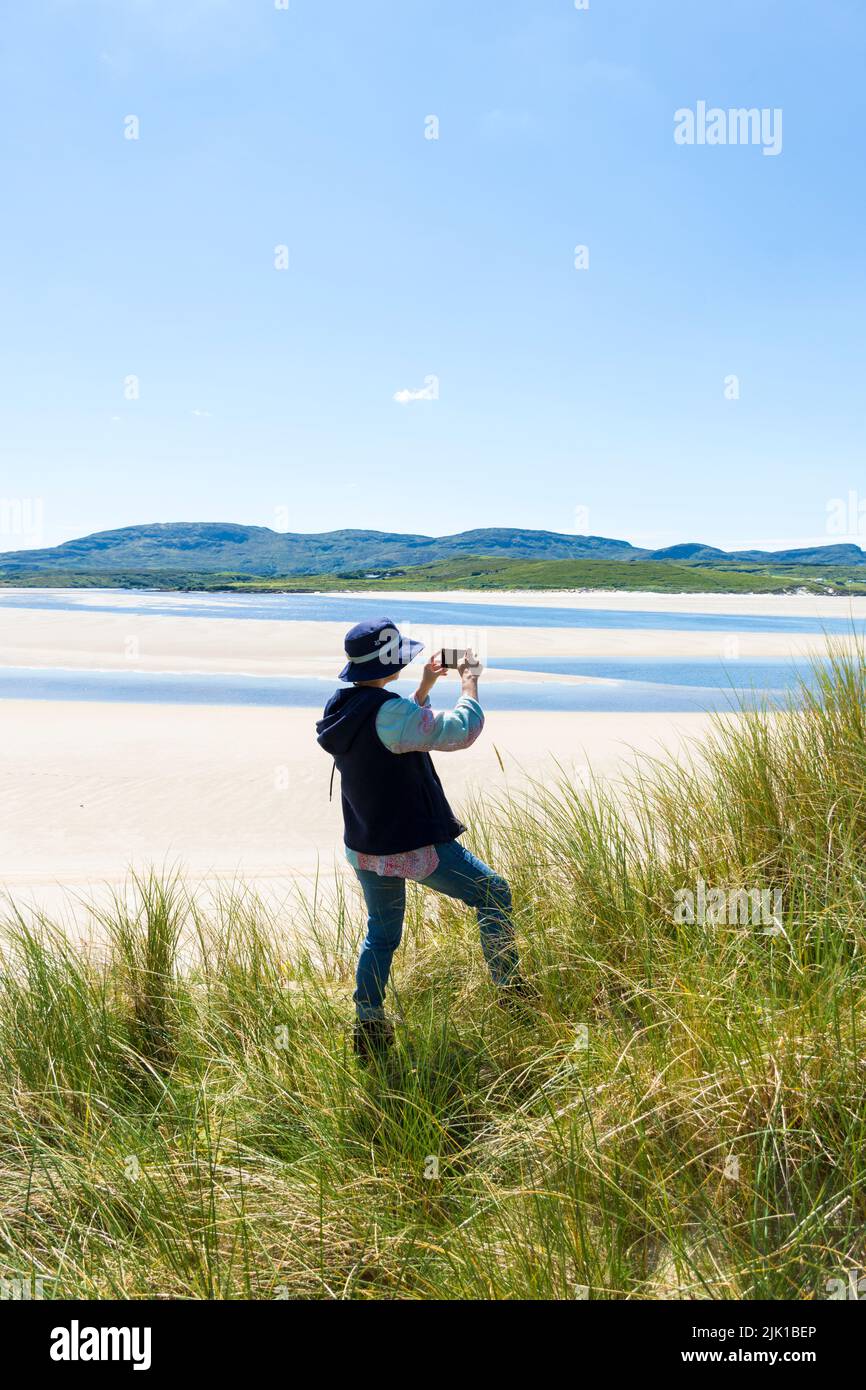 Mujer toma una foto con la cámara del teléfono en Ballinreavy Strand junto a la Reserva Natural Sheskinmore. Sheskinmore se refiere a una gran área de dunas de arena, el lago An Foto de stock