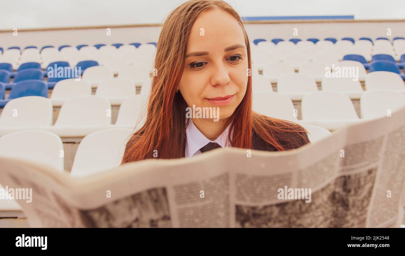 Una mujer en un traje de negocios lee un periódico mientras está sentada en el estadio. Foto de stock