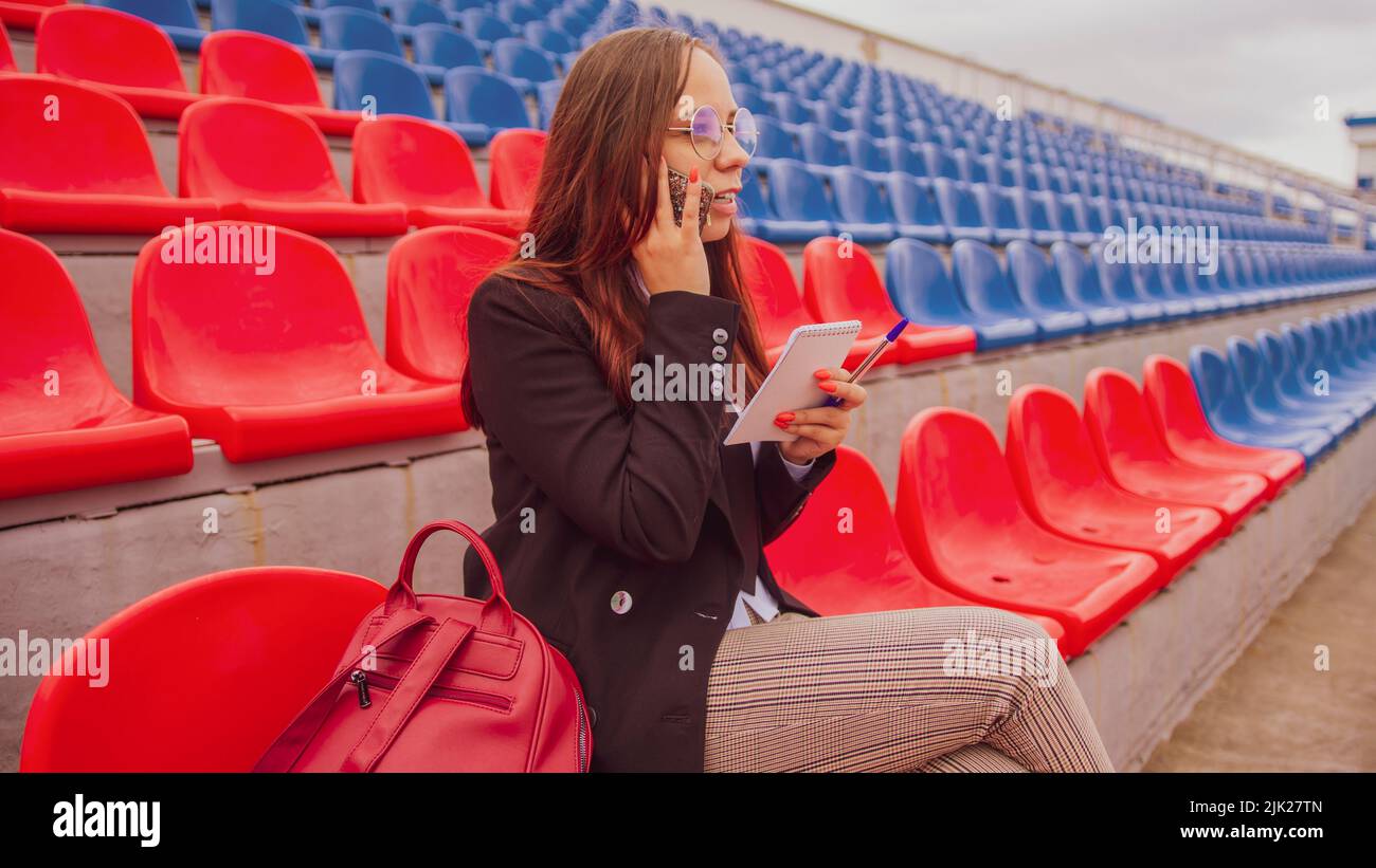 Mujer joven en vasos con bloc de notas, bolígrafo hablando en el teléfono móvil, sentado en gradas estadio. Periodista hablando en smartphone, hablando c Foto de stock