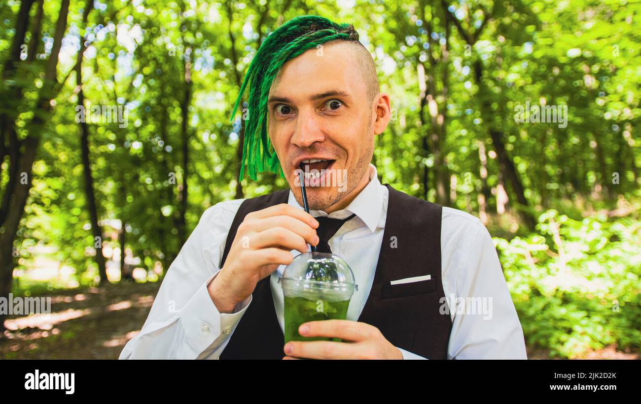 Joven con temblos bebiendo limonada refrescante en el parque forestal. Hipster divertido con peinado verde beber refresco a través de la paja, apagando Foto de stock