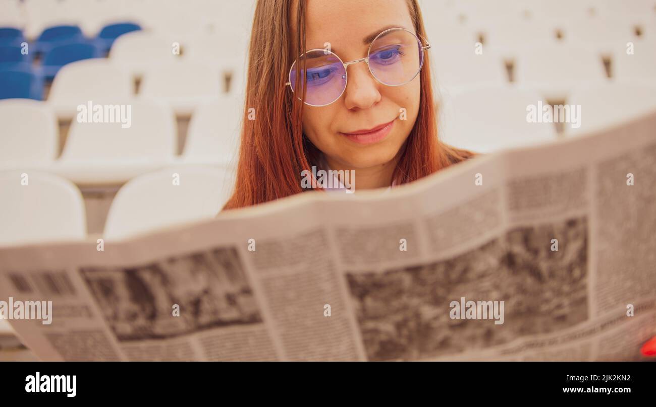 Una mujer con gafas y un traje de negocios lee un periódico mientras está sentada en el estadio. Foto de stock