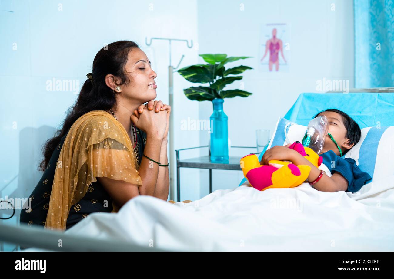 Madre preocupada orando por la hija enferma para la recuperación de la salud en el hospital cerca de la cama - concepto de creencia emocional, cuidado familiar y paternidad. Foto de stock