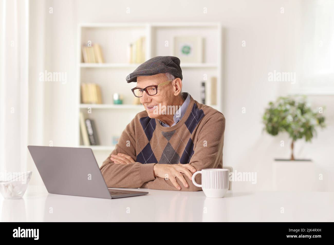 Hombre anciano feliz sentado en un escritorio en casa y mirando a un ordenador portátil Foto de stock