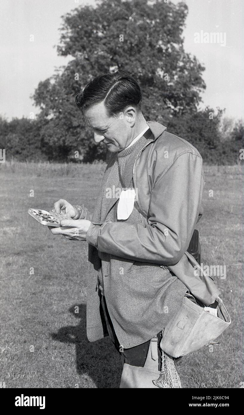 1960s, histórico, al aire libre en el campo, un pescador grueso, llevando una pequeña bandeja en busca de un gancho, Inglaterra, Reino Unido. Ropa de pesca de la época, bolsa de lona para el hombro, chaqueta impermeable y suéter sobre chaqueta deportiva. Foto de stock