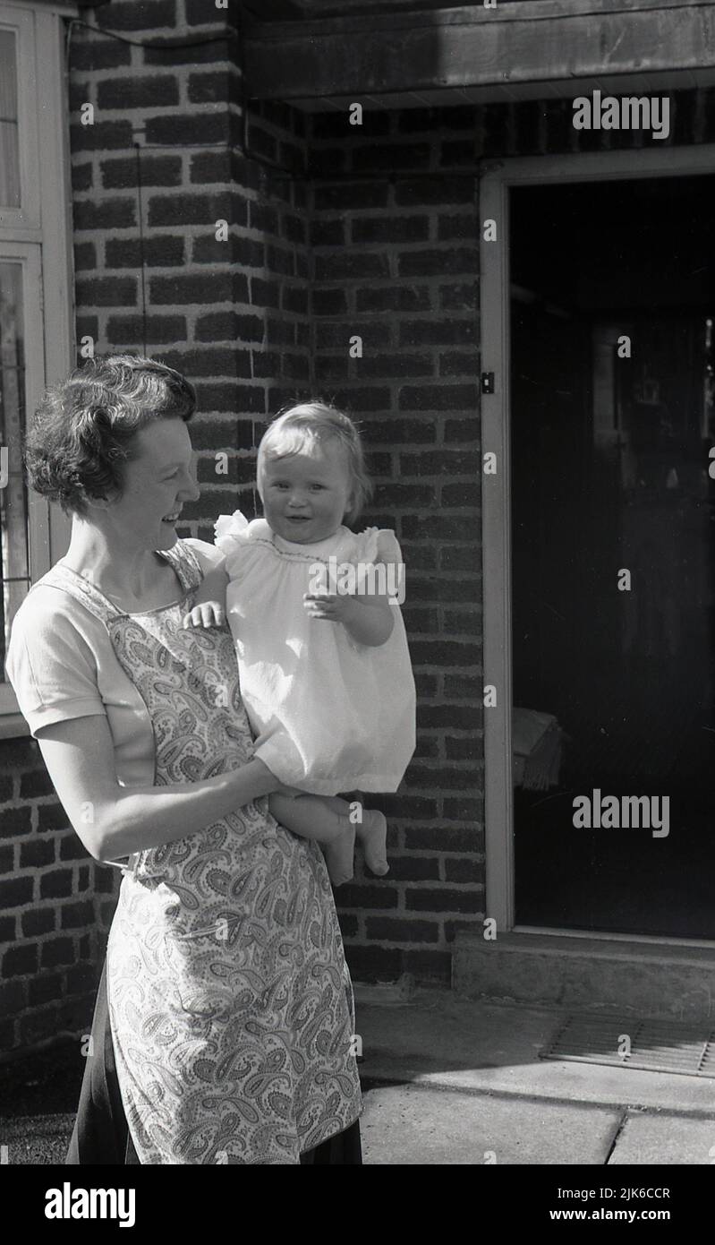 1960s, histórico, fuera de la entrada de una casa, una madre que llevaba un pinafore estampado de la época, sosteniendo a su hija infantil, Inglaterra, Reino Unido. Foto de stock