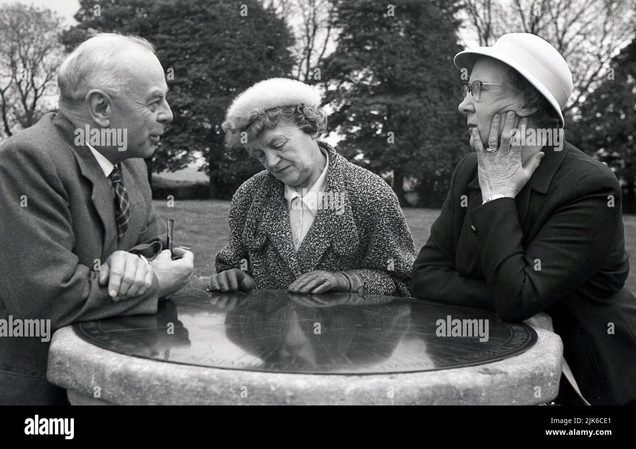 1960s, histórico, fuera en un parque, tres ancianos bien vestidos apoyándose en la cara de bronce de un reloj de sol de piedra, charlando, Inglaterra, Reino Unido. Foto de stock