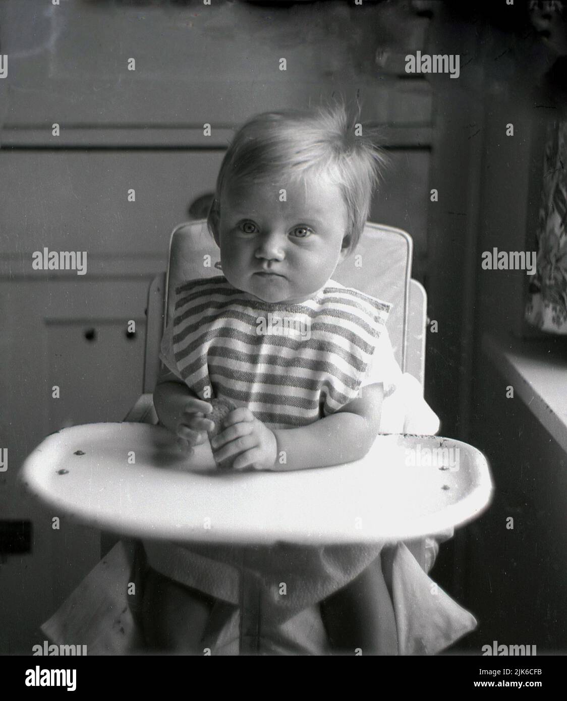 1960s, histórico, niño sentado en una silla alta, con una bandeja de metal esmaltado, sosteniendo una galleta, Inglaterra, REINO UNIDO. Foto de stock