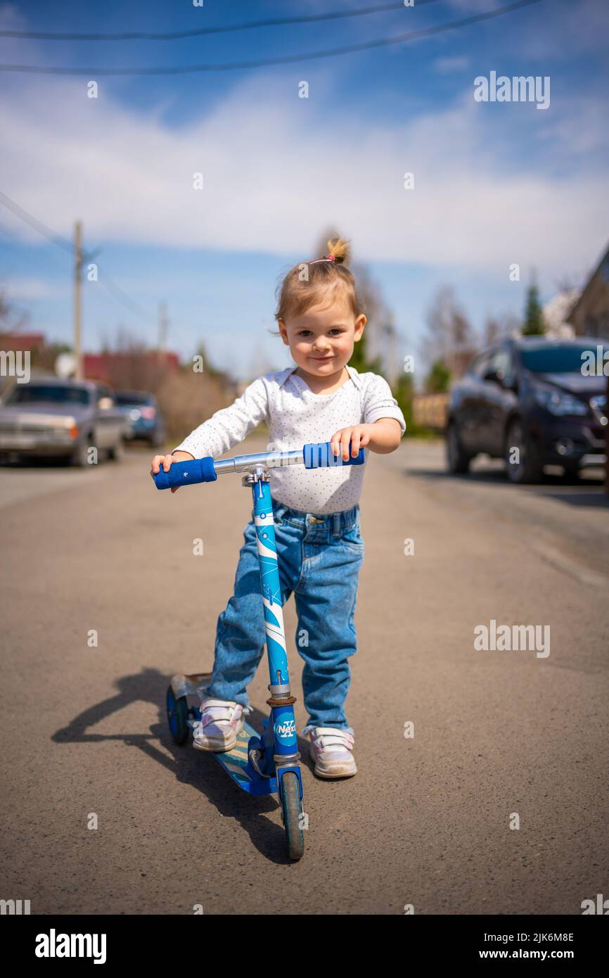 Linda niña pequeña en monos azules montando en scooter de patada. Feliz sano niño pequeño encantador que se divierte en la ciudad. Niño activo en el día frío Foto de stock