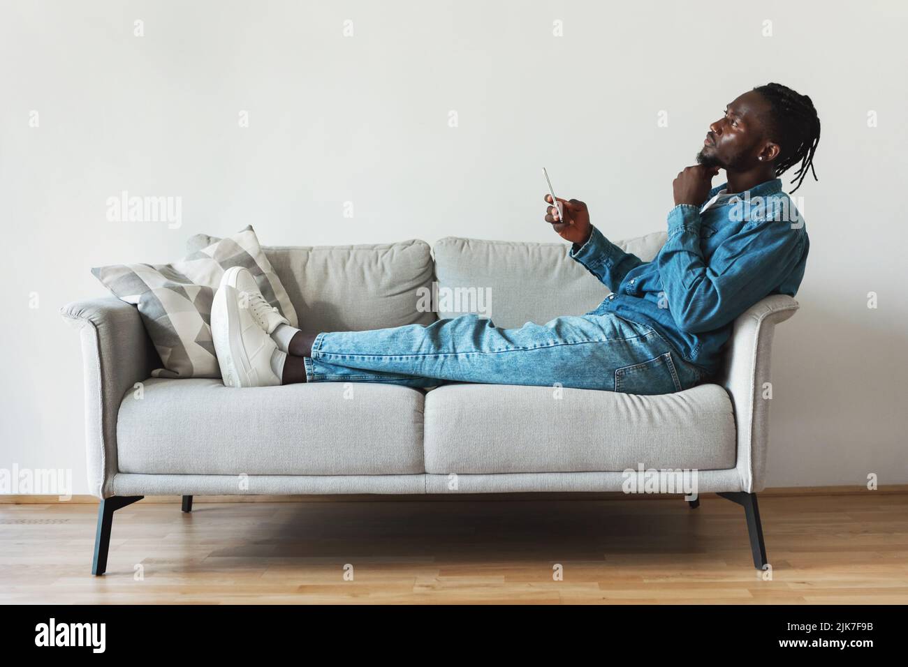 Hombre negro pensativo usando el teléfono móvil sentado en el sofá interior Foto de stock