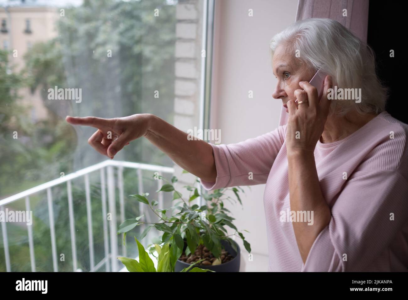 Mujer mayor hablando por teléfono escuchando y escuchando rumores o chismes de pie en casa cerca de la ventana Foto de stock
