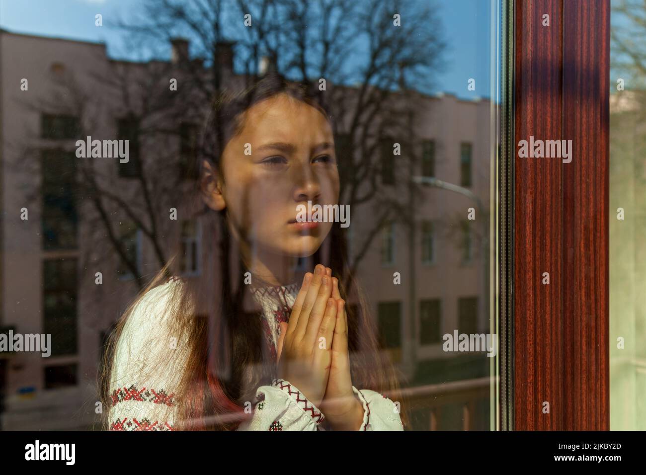 Una chica con un vestido bordado reza cerca de la ventana. La niña protesta contra la guerra y muestra patriotismo y amor por Ucrania. Ucraniano-ruso Foto de stock