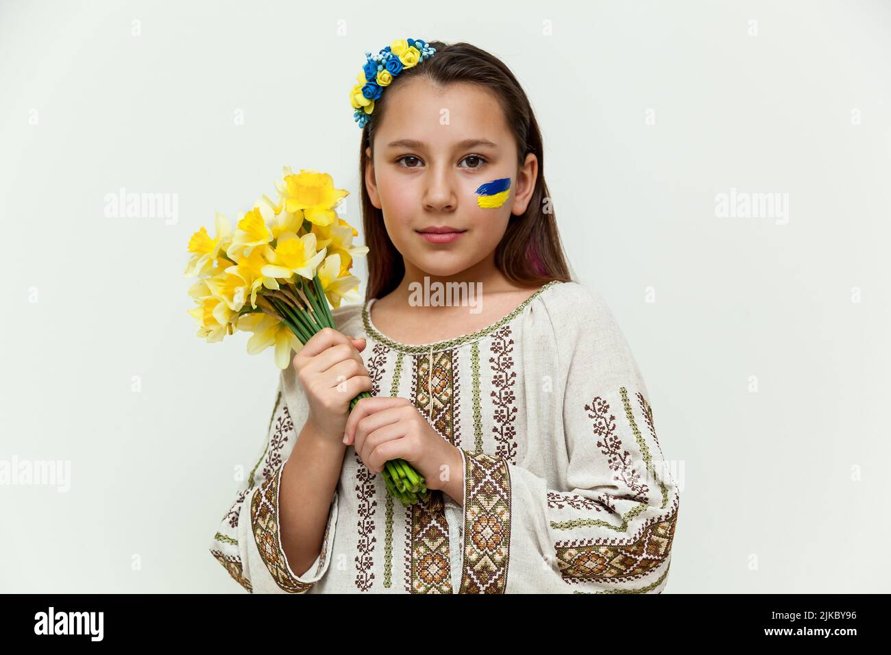 Una niña con un vestido bordado se coloca contra una pared blanca, sosteniendo un ramo de narcisos amarillos en sus manos. Foto de stock