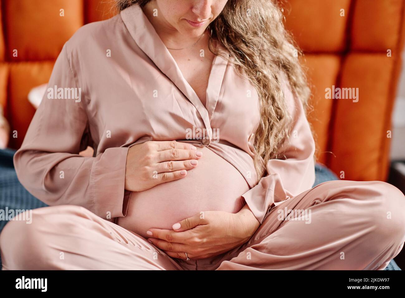 Joven mujer embarazada en ropa de hogar rosa tocando su barriguita con el futuro bebé mientras se sienta en la cama y se prepara para dormir Foto de stock