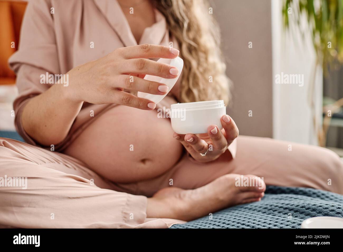 Manos de una joven embarazada sosteniendo un frasco blanco de plástico abierto y su cubierta mientras va a aplicar crema en sus manos o cuerpo Foto de stock