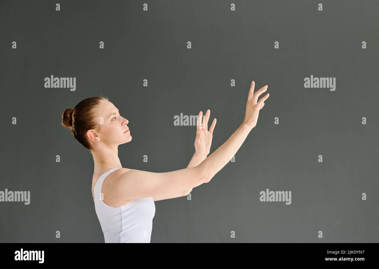 Joven bailarín de ballet con atuendo blanco manteniendo los brazos levantados durante el ejercicio o la danza en el escenario o en el aula sobre fondo gris Foto de stock