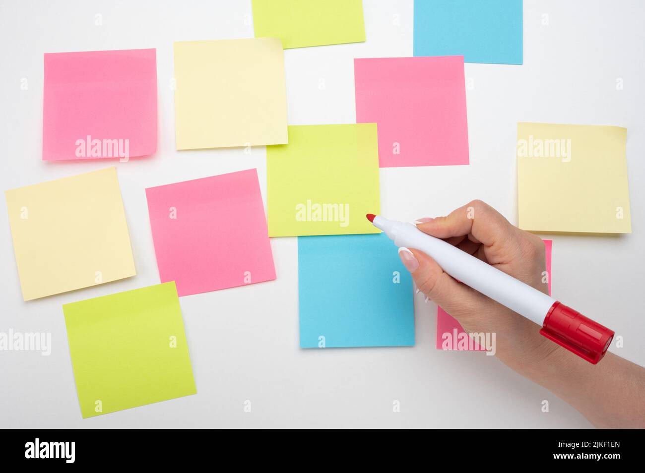 Muchas notas adhesivas vacías de color en la pared blanca. Mano con marcador rojo listo para escribir. Notas adhesivas en blanco sobre fondo blanco. Papel de notas adhesivo para maqueta Foto de stock