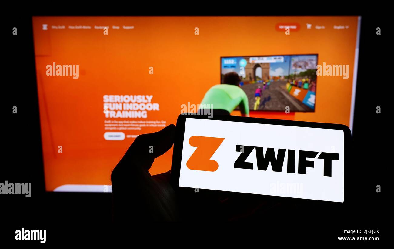 Persona que sostiene el teléfono móvil con el logotipo de la compañía americana de juegos de fitness Zwift Inc. En la pantalla delante de la página web. Enfoque la pantalla del teléfono. Foto de stock