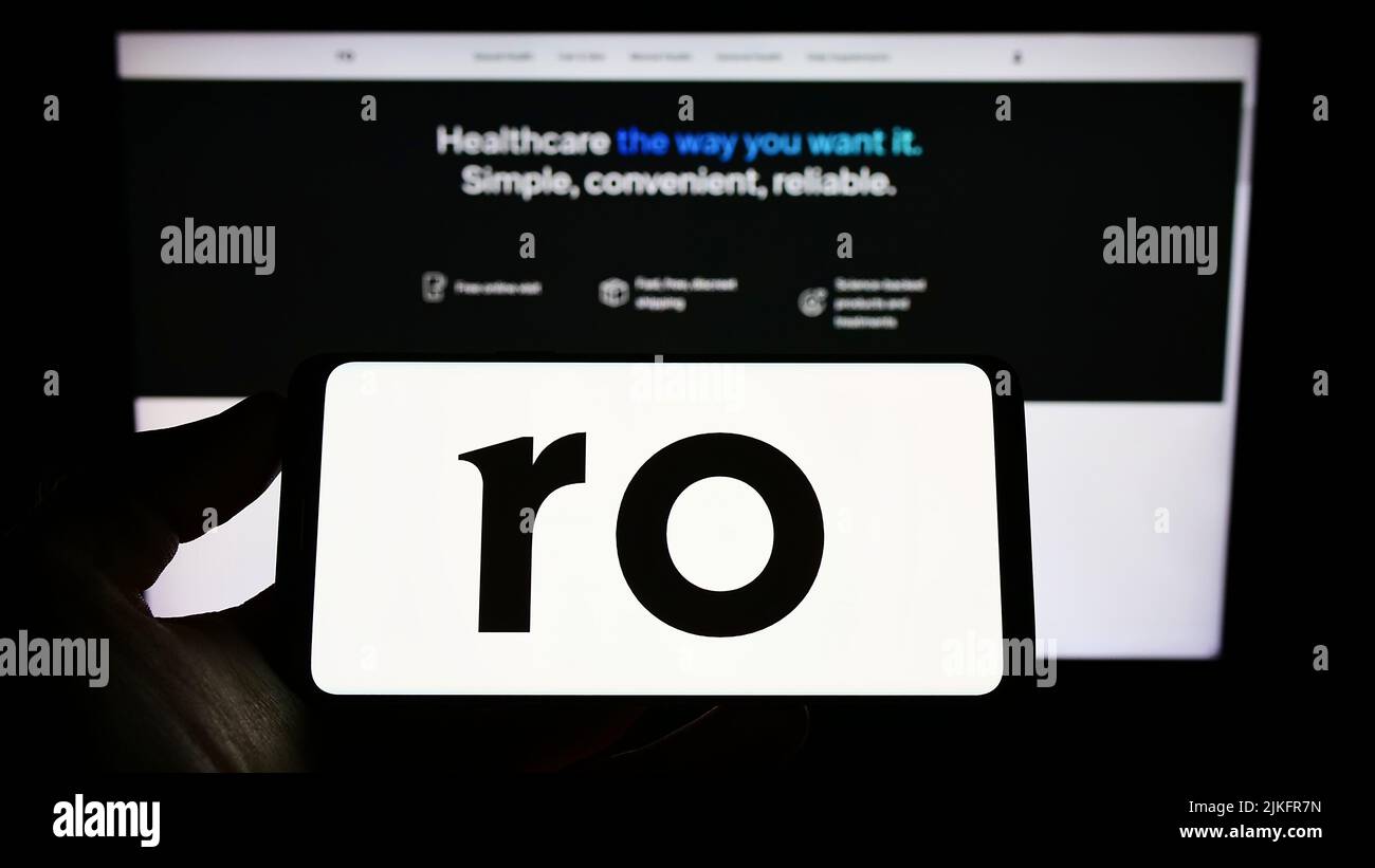 Persona que sostiene smartphone con el logotipo de la empresa de telesalud Roman Health Ventures Inc. (RO) en la pantalla delante del sitio web. Enfoque la pantalla del teléfono. Foto de stock