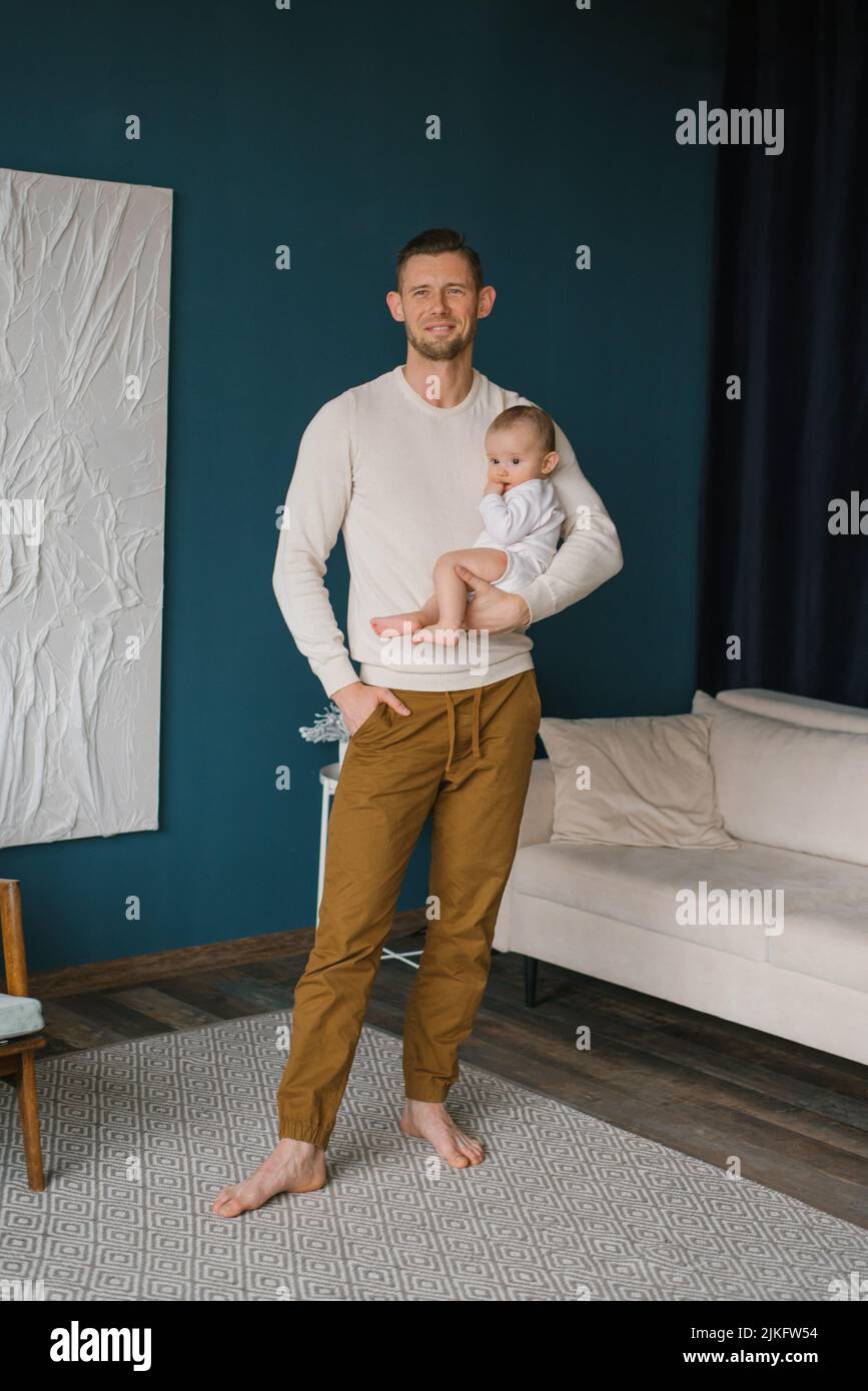 El joven es feliz y sostiene a su hijo de seis meses en sus brazos en su casa Foto de stock